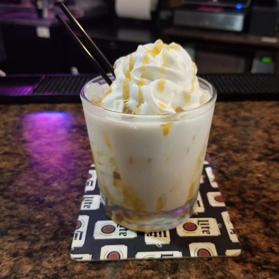 A creamy, caramel-drizzled drink with whipped cream and black straws on a branded coaster at Mel's Roadhouse.