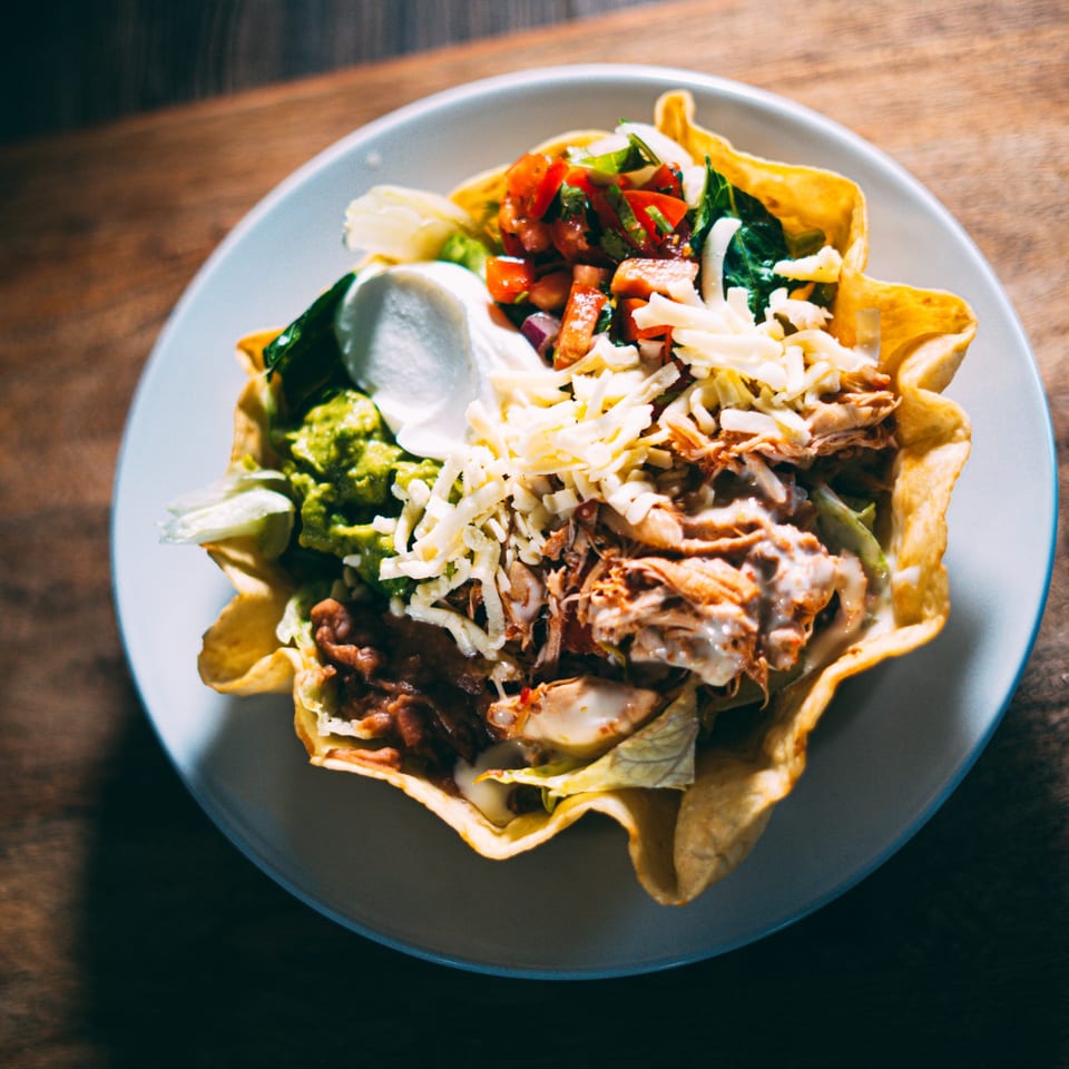 A taco salad in a crispy tortilla bowl, topped with shredded meat, cheese, guacamole, and salsa.