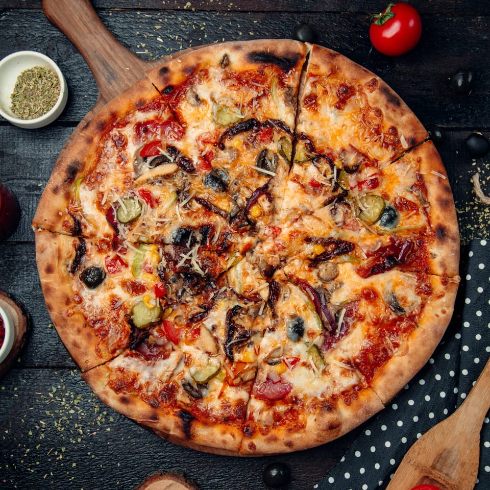A sliced Mel's Roadhouse pizza with various toppings, including olives, peppers, and cheese, on a wooden board.