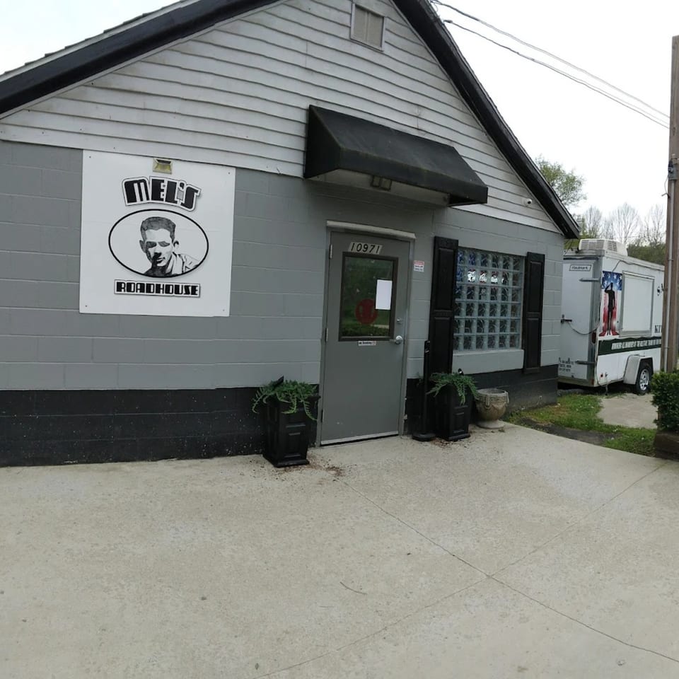 Exterior of Mel's Roadhouse, a gray building with a black awning and a sign featuring a man's portrait.