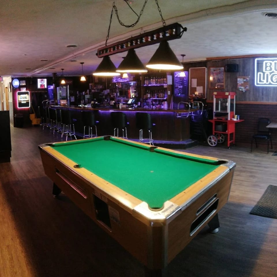 A pool table in the foreground of Mel's Roadhouse bar with purple lighting and a popcorn machine.